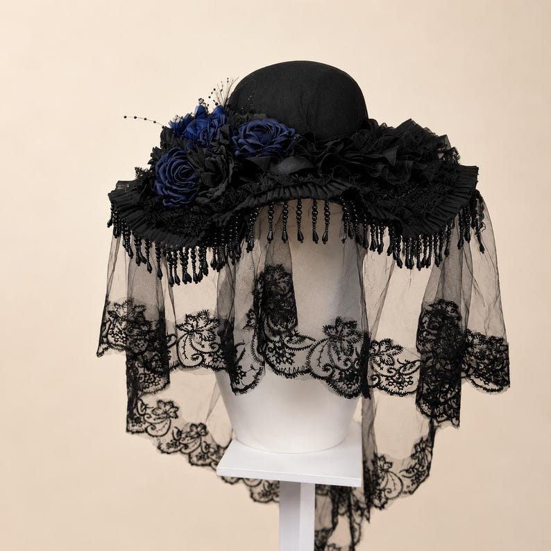 Puede incluir: Un sombrero negro con ala ancha adornado con rosas de tela azul oscuro y negras, ribete de volantes negros y flecos de cuentas negras colgantes. Un velo largo de encaje negro transparente con bordados florales cuelga del ala.