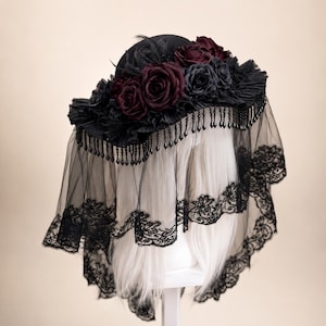 Puede incluir: Un sombrero negro adornado con rosas de tela en color burdeos y negro, plumas negras y un ala plisada. Un largo velo de tul negro con un borde de encaje festoneado cuelga del ala, acentuado por un fleco de peque&ntilde;as cuentas negras en forma de l&aacute;grima.