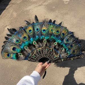 May include: A handheld folding fan featuring iridescent peacock feathers in shades of blue, green, and gold. The base is constructed from black material with intricate gold patterns, accented by dangling gold chains and green teardrop-shaped gems.