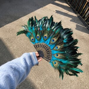 May include: A handheld folding fan featuring vibrant teal and dark green peacock feathers arranged in a semi-circular pattern. The base of the fan is constructed from black material with intricate gold-colored filigree detailing along the ribs.