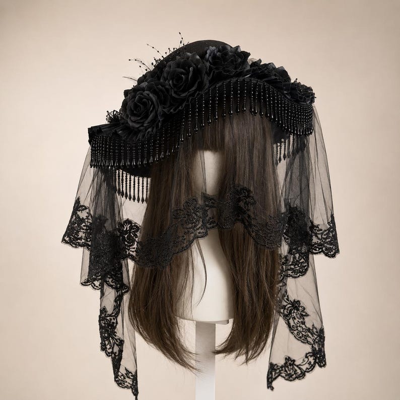 Puede incluir: Un sombrero negro adornado con un grupo de rosas de tela negra a lo largo de la corona. El ala presenta un borde de flecos de cuentas negras colgantes. Un velo negro largo y transparente con intrincados bordados de encaje floral en los bordes.