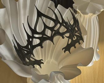 Peut inclure: Une couronne noire complexe et pointue avec un design en treillis acéré. La couronne est placée à l'intérieur d'un grand objet blanc en forme de trompette avec des plis verticaux profonds, à côté d'un objet cylindrique sombre et cannelé.
