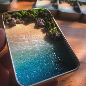 Könnte beinhalten: Eine rechteckige Metalldose mit einem Miniatur-Diorama einer Küstenlandschaft. Die Szene zeigt einen Sandstrand, weiße Wellen aus Kunstharz und tiefblaues Meerwasser. Dunkelgraue Felsen und grüne Moosflecken säumen den Rand des Sandstrandes.