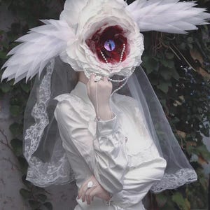 Op de afbeelding: Een witte jurk met lange mouwen, ruches op de schouders en een bijpassend hoofddeksel met een grote witte bloem met een donkerrood hart, een paars oog en witte veerachtige accenten. Een witte kanten sluier hangt aan het hoofddeksel.