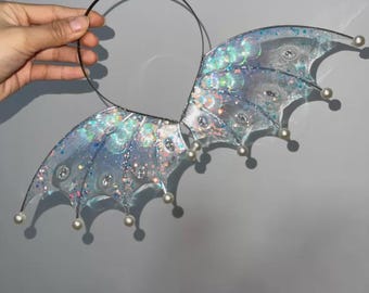 May include: A headband featuring two translucent, iridescent wing-shaped attachments. The wings have a blue and silver color palette with embedded glitter, clear faceted accents, and small white spherical beads at the tips of each wing segment.