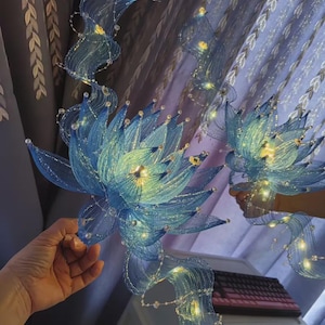 Könnte beinhalten: Zwei blaue Lotusblütenskulpturen aus feinem Drahtgeflecht mit geschichteten Blütenblättern und langen, fließenden Bändern. Jede Blume wird von kleinen, warmweißen LED-Lichterketten beleuchtet und ist mit winzigen, klaren Perlen an den Rändern verziert.