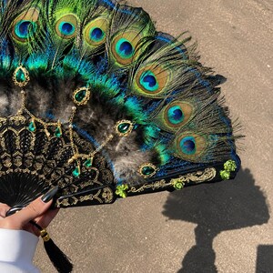 May include: A handheld folding fan featuring iridescent peacock feathers in shades of green and blue. The black frame has intricate gold filigree, accented with dangling emerald-colored teardrop gems, small green floral embellishments, and a black tassel.
