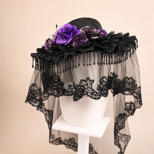 Puede incluir: Un sombrero negro de ala ancha adornado con rosas de tela de color p&uacute;rpura intenso y flores oscuras m&aacute;s peque&ntilde;as. El ala presenta un borde de tela negra con volantes, cuentas negras en forma de l&aacute;grima y un largo velo de encaje negro transparente.