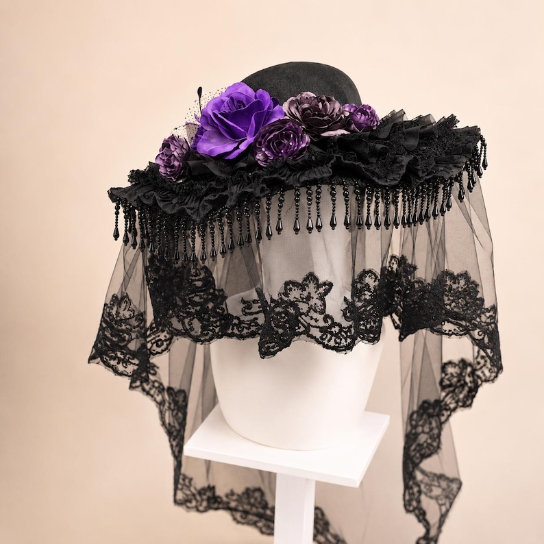 Puede incluir: Un sombrero negro de ala ancha adornado con rosas de tela de color p&uacute;rpura intenso y flores oscuras m&aacute;s peque&ntilde;as. El ala presenta un borde de tela negra con volantes, cuentas negras en forma de l&aacute;grima y un largo velo de encaje negro transparente.