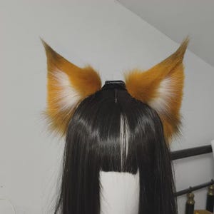 May include: A pair of pointed, faux fur animal ears in shades of orange, brown, and white, attached to a black headband. The ears feature a dense, textured appearance with white fur detailing on the inner sections, positioned atop a black wig.