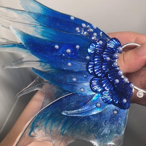 May include: A decorative ear cuff shaped like a wing, featuring translucent blue and white resin with glitter accents. The wing is adorned with small white pearls and layered, metallic blue scale-like elements near the silver-toned metal ring.