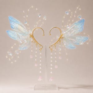 May include: A pair of gold-tone ear cuffs featuring iridescent blue wing-shaped attachments, delicate wire tendrils with small clear beads, and dangling chains adorned with white pearls and small pink floral-shaped beads on a clear stand.