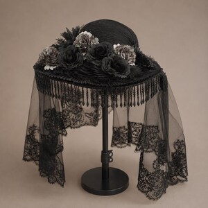Puede incluir: Un sombrero negro con ala ancha adornado con un grupo de rosas de tela oscura y follaje. El borde del ala est&aacute; decorado con flecos de cuentas negras colgantes y cuelga un largo velo de encaje negro transparente con patrones florales.