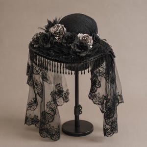 Puede incluir: Un sombrero negro con ala ancha adornado con un grupo de rosas de tela oscura y ribete con volantes. El sombrero est&aacute; decorado con un largo velo de encaje negro transparente que cuelga del ala, acentuado por una fila de flecos de cuentas negras.
