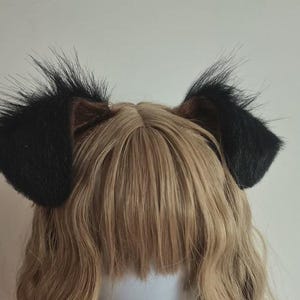 Peut inclure: Une paire d'oreilles d'animal en fausse fourrure noire fixée sur un serre-tête, avec de longues fibres synthétiques noires hérissées sur les bords extérieurs et une doublure intérieure marron foncé lisse, posée sur des cheveux synthétiques blonds ondulés.