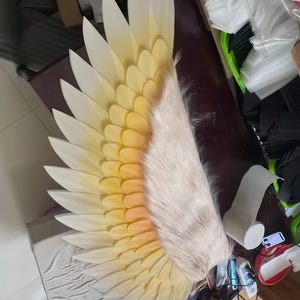 May include: A large, single decorative wing featuring layered, pointed white feathers that transition into a soft yellow gradient toward the base. The inner section is covered in textured, off-white faux fur, creating a realistic appearance.