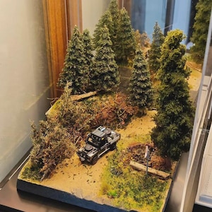 May include: A miniature diorama featuring a black off-road truck model parked on a sandy dirt path. The scene includes several tall green pine trees, scattered brown logs, and textured ground cover, all enclosed within a clear display case.