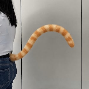 Könnte beinhalten: Ein langer, plüschiger Katzen-Schwanz-Aufsatz mit orangefarbenen und cremefarbenen Streifen. Das Accessoire hat eine weiche, pelzige Textur mit abwechselnden horizontalen Bändern in Hellorange und Weiß, die sich vom Hosenbund einer blauen Jeans abheben.