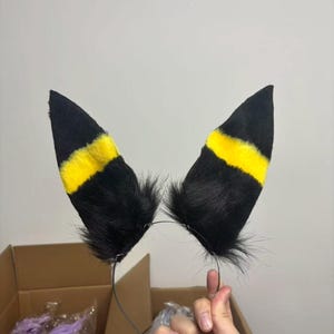 Puede incluir: Una diadema con dos orejas altas y puntiagudas hechas de piel sintética negra. Cada oreja tiene una franja horizontal de color amarillo brillante en la sección central. Las orejas están unidas a una diadema de metal fino de color oscuro.