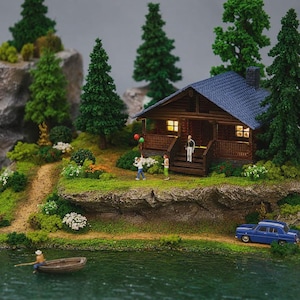 May include: A miniature diorama featuring a brown log cabin with a blue roof, illuminated windows, and a porch. The scene includes green pine trees, rocky terrain, a small blue car, and a person in a rowboat on a body of water with figures.