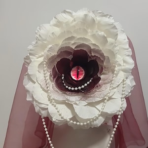 May include: A large white paper flower headpiece featuring a dark burgundy centre with a central red reptilian eye. The flower is adorned with a draped string of white faux pearls and is attached to a sheer burgundy veil that hangs behind it.