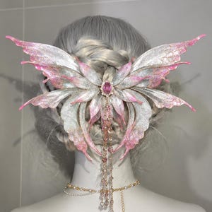 May include: A hair accessory featuring large, iridescent butterfly wings in shades of silver and pink. The center is adorned with a single oval pink gemstone, from which delicate gold chains with small pink beads hang down over the hair.