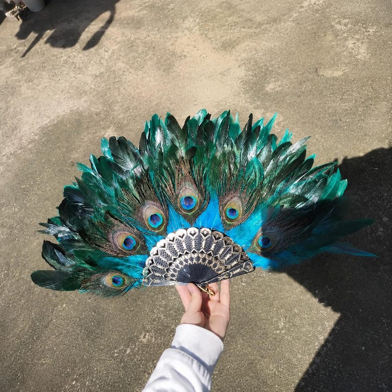 May include: A handheld folding fan featuring vibrant teal and dark green peacock feathers arranged in a semi-circular pattern. The base of the fan is constructed from dark, intricate, laser-cut material with a metallic gold ring at the pivot point.