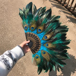 May include: A handheld folding fan featuring vibrant teal and dark green peacock feathers arranged in a semi-circular pattern. The base of the fan is constructed from black material with intricate gold-colored filigree detailing throughout.