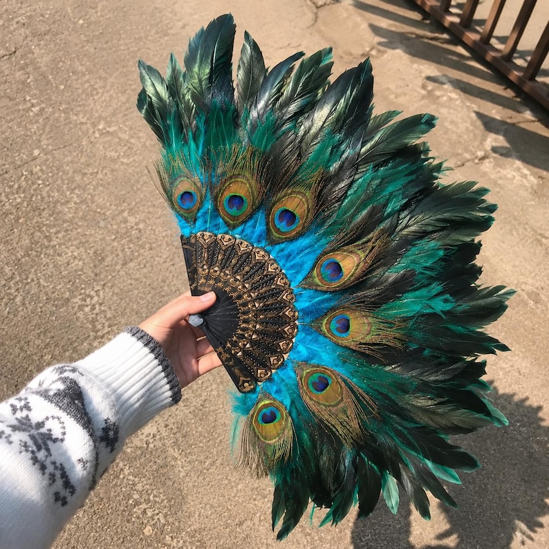 May include: A handheld folding fan featuring vibrant teal and dark green peacock feathers arranged in a semi-circular pattern. The base of the fan is constructed from black material with intricate gold-colored filigree detailing throughout.