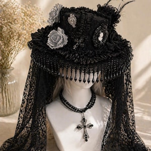 Puede incluir: Un sombrero gótico negro con ala ancha, adornado con rosas plateadas, un camafeo y flecos de cuentas. Un velo de encaje negro cae, combinado con un collar de cuentas negro con un colgante de cruz. El sombrero se exhibe en una cabeza de maniquí blanca.