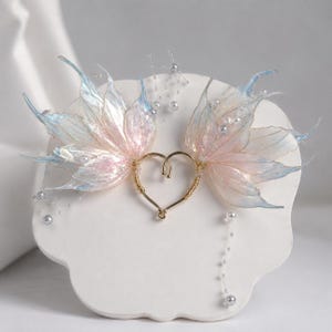 May include: An ear cuff featuring a gold-toned heart-shaped wire frame adorned with iridescent, translucent wing-like structures in shades of pale pink and light blue, accented with small white beads and delicate dangling strands of tiny white pearls.