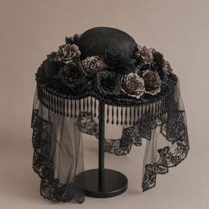 Puede incluir: Un sombrero negro adornado con un denso arreglo de flores de tela oscura alrededor de la copa. El ala presenta un fleco colgante de cuentas negras y un largo velo de encaje negro transparente que cae desde los bordes de la estructura circular.