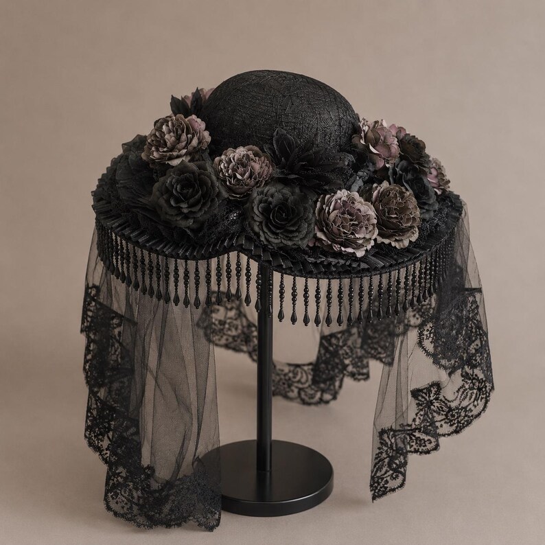 Puede incluir: Un sombrero negro adornado con un denso arreglo de flores de tela oscura alrededor de la copa. El ala presenta un fleco colgante de cuentas negras y un largo velo de encaje negro transparente que cae desde los bordes de la estructura circular.