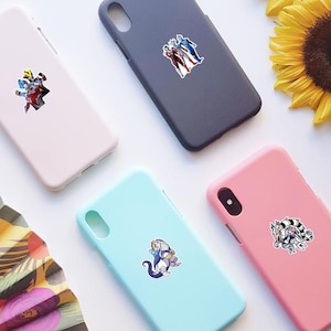 Puede incluir: Cuatro fundas para smartphone de colores s&oacute;lidos en rosa claro, gris oscuro, azul claro y rosa brillante. Cada funda presenta una pegatina &uacute;nica de un personaje de la serie Ultraman, incluyendo figuras humanoides y un dise&ntilde;o de monstruo.