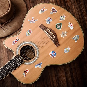 Puede incluir: Un cuerpo de guitarra ac&uacute;stica de color claro decorado con numerosas pegatinas peque&ntilde;as y coloridas que muestran varios personajes y logotipos de Ultraman. La guitarra tiene un puente de madera oscura, una boca circular y un m&aacute;stil.