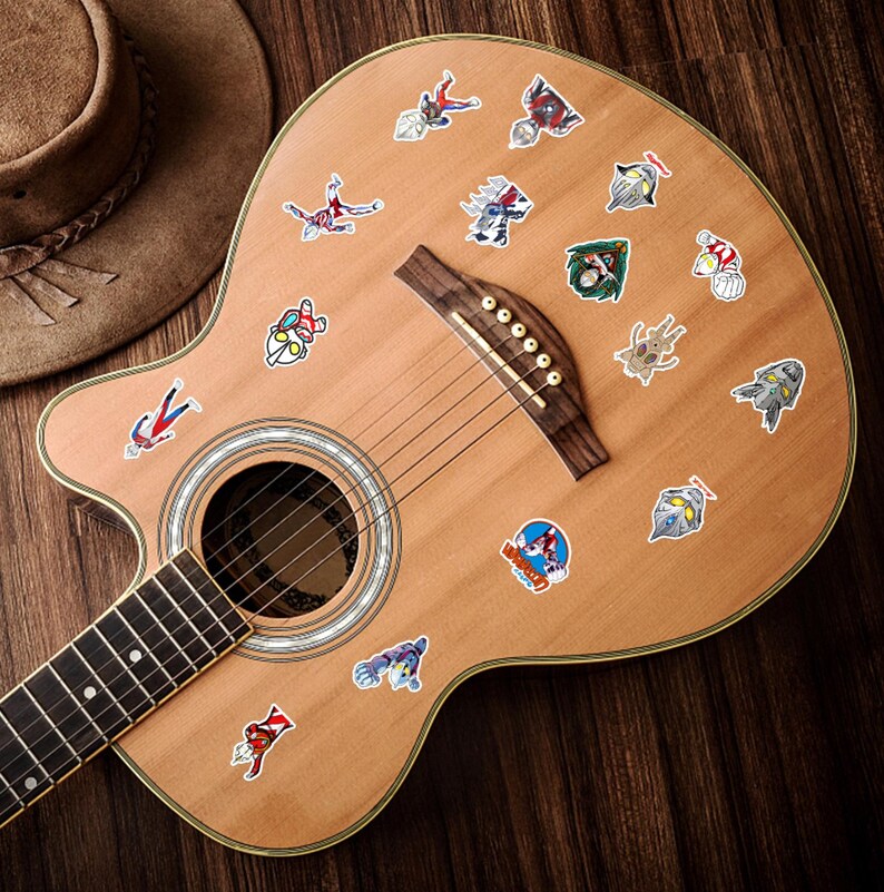 Puede incluir: Un cuerpo de guitarra ac&uacute;stica de color claro decorado con numerosas pegatinas peque&ntilde;as y coloridas que muestran varios personajes y logotipos de Ultraman. La guitarra tiene un puente de madera oscura, una boca circular y un m&aacute;stil.
