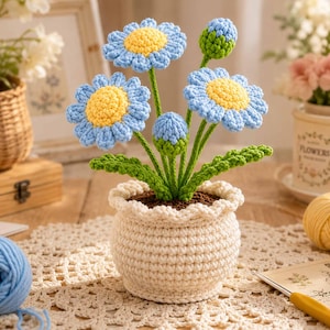 May include: A crochet potted plant featuring two fully bloomed light blue daisies with bright yellow centers and two closed flower buds. The flowers have green stems and leaves, all housed in a textured, cream-colored crocheted pot with a ruffled rim.