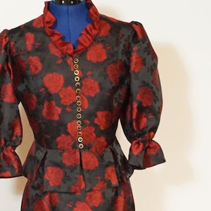 Puede incluir: Un vestido negro y rojo con estampado floral. El vestido tiene un corpiño ajustado con un peplum y una falda que llega a la rodilla. El vestido tiene un cuello con volantes y mangas cortas con volantes en los puños.