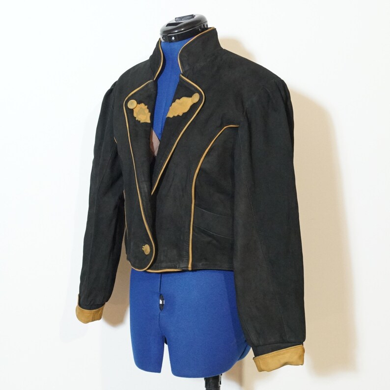 May include: A black cropped leather jacket with a brown trim. The jacket has a stand-up collar and a single button closure. The jacket has a slightly puffed shoulder design.