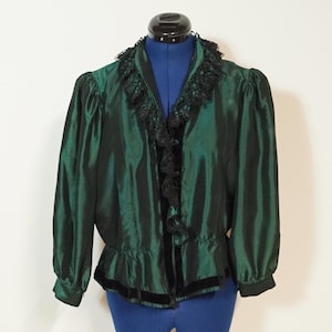 Puede incluir: Una blusa de manga larga en color verde esmeralda oscuro con un cuello de volantes de encaje negro y ribete a lo largo de la abertura frontal. La prenda tiene un acabado ligeramente iridiscente, hombros fruncidos y una banda de terciopelo.