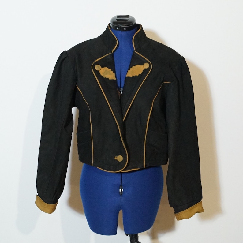 May include: A black cropped jacket with a brown trim and gold buttons. The jacket has a stand-up collar and long sleeves.