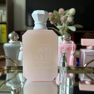 May include: A light pink perfume bottle with a white geometric cap and the text "KAY ALI" and "YUM DOUBLE MARSHMALLOW | 61". A small, clear glass atomizer is in front of the bottle. Other perfume bottles are in the background.