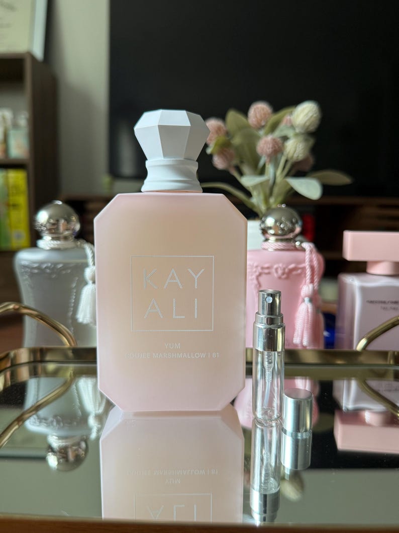 May include: A light pink perfume bottle with a white geometric cap and the text "KAY ALI" and "YUM DOUBLE MARSHMALLOW | 61". A small, clear glass atomizer is in front of the bottle. Other perfume bottles are in the background.