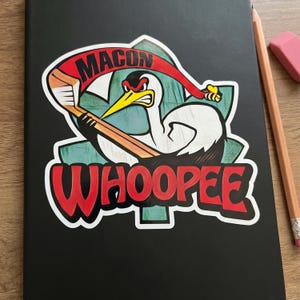 May include: A black notebook featuring a sticker with the text "MACON" on a red banner and "WHOOPEE" in red. The sticker depicts a cartoon bird holding a hockey stick. A pencil and eraser are visible.