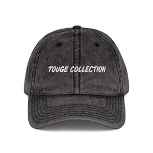 May include: A charcoal gray baseball cap with the text "TOUGE COLLECTION" embroidered in white across the front. The cap has a curved brim and a distressed, vintage look.