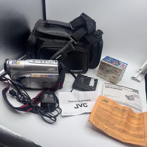 Puede incluir: Una videocámara JVC Compact VHS plateada con lente de zoom 700x, acompañada de un bolso de transporte negro, un cargador de batería, tres cintas JVC VHS-C, un manual de JVC, una tarjeta de garantía y una nota de agradecimiento de JVC.