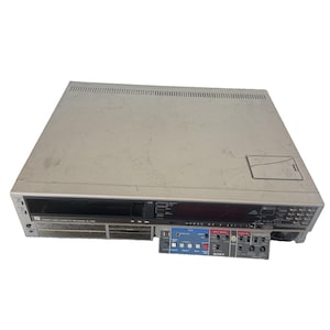 May include: A silver Sony SL-2700 stereo video cassette recorder with a top-loading mechanism. The front panel features a black display area, various control buttons, and a fold-down door revealing additional settings and input controls.