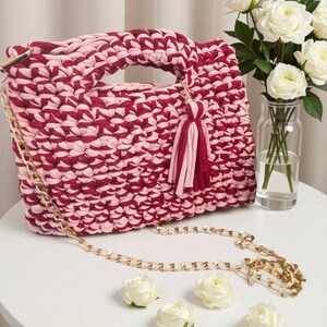 K&ouml;nnte beinhalten: Handgefertigte, zweifarbige rosa-burgunderfarbene H&auml;keltasche mit goldener Kette und Quaste. Die Tasche hat einen Griff und steht auf einem wei&szlig;en Tisch mit wei&szlig;en Rosen.