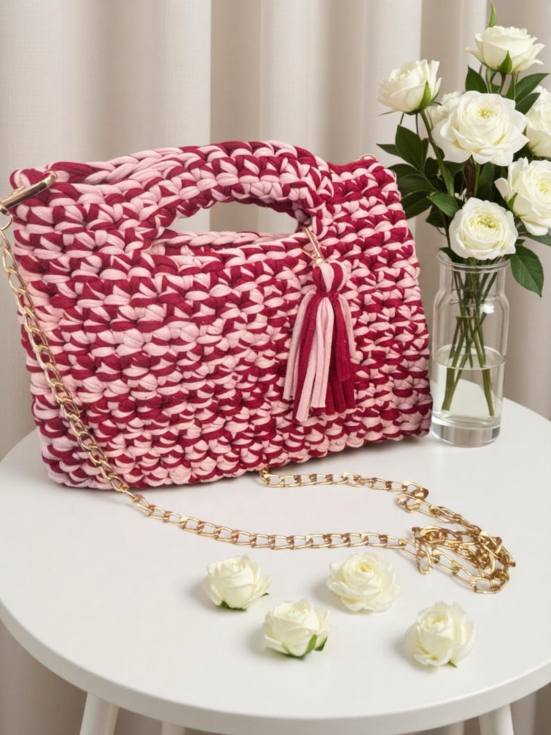 K&ouml;nnte beinhalten: Handgefertigte, zweifarbige rosa-burgunderfarbene H&auml;keltasche mit goldener Kette und Quaste. Die Tasche hat einen Griff und steht auf einem wei&szlig;en Tisch mit wei&szlig;en Rosen.