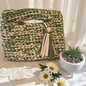 K&ouml;nnte beinhalten: Handgefertigte gr&uuml;ne und cremefarbene H&auml;keltasche mit rechteckiger Form und Griff. Die Tasche hat einen goldfarbenen Kettenriemen und einen Quastenanh&auml;nger. G&auml;nsebl&uuml;mchen und eine kleine Sukkulente in einem wei&szlig;en Topf sind ebenfalls im Bild.
